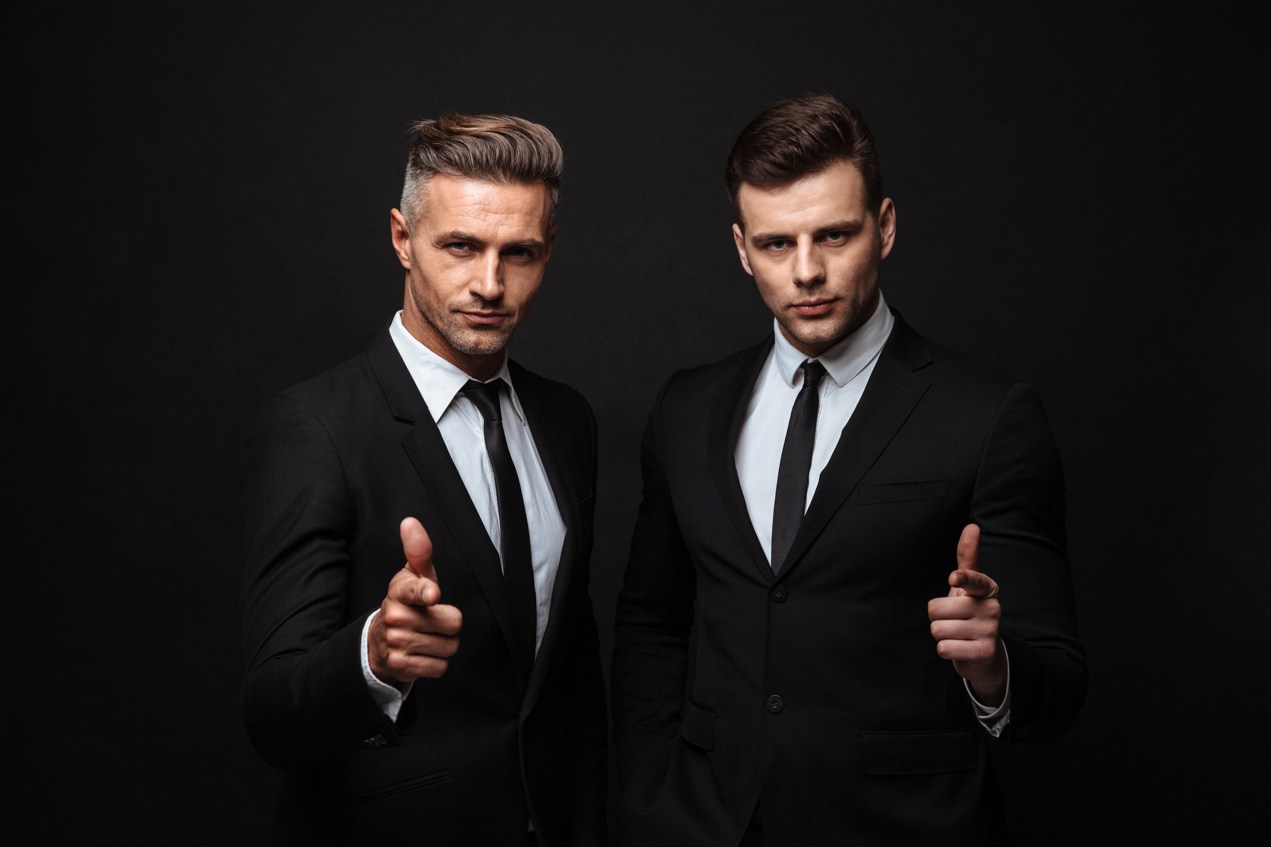 two-confident-handsome-businessmen-wearing-suit-standing-isolated-black-wall-pointing-camera.jpg